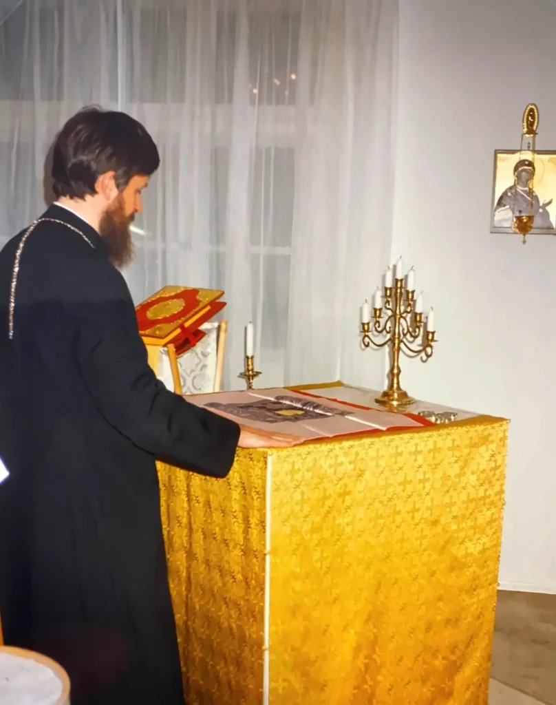 Vater Peter in schwarzem Gewand steht am golden verhüllten Altartisch und legt die Hand auf das ausgebreitete Antimension, daneben ein siebenarmiger Leuchter, an der Wand eine Ikone