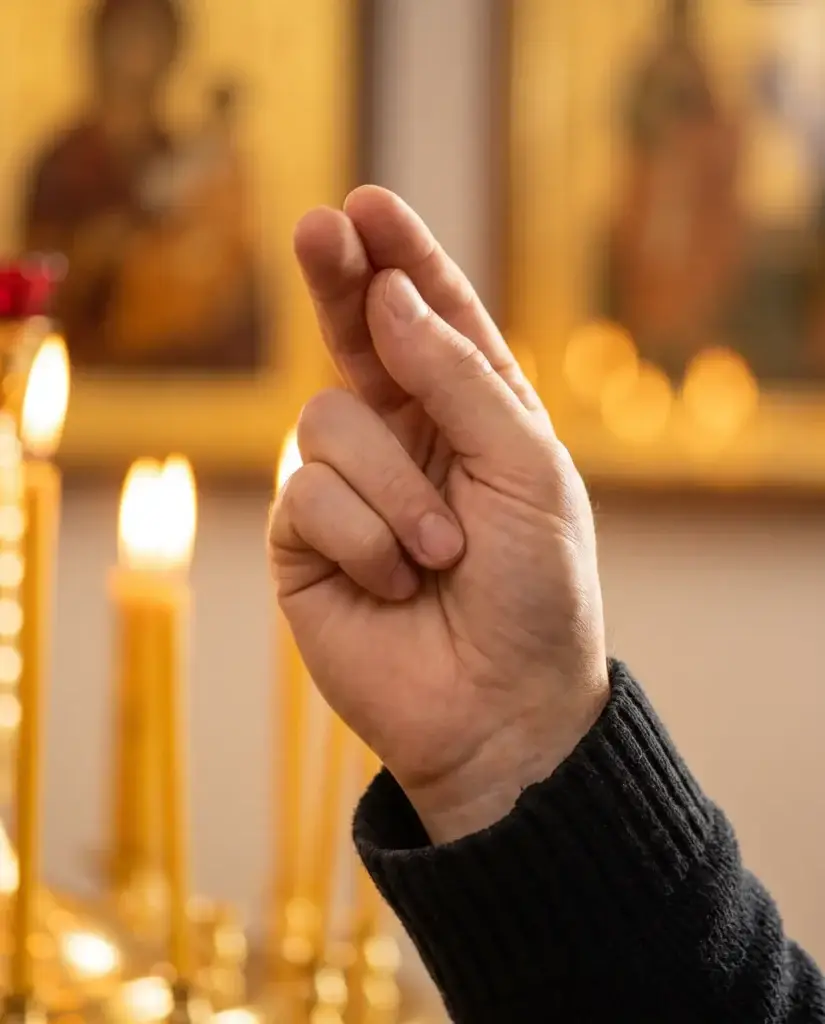 Rechte Hand beim Kreuzzeichen: Daumen, Zeige- und Mittelfinger zusammengelegt, Ringfinger und kleiner Finger an der Handfläche, dahinter unscharf eine Ikone und Kerzenlicht
