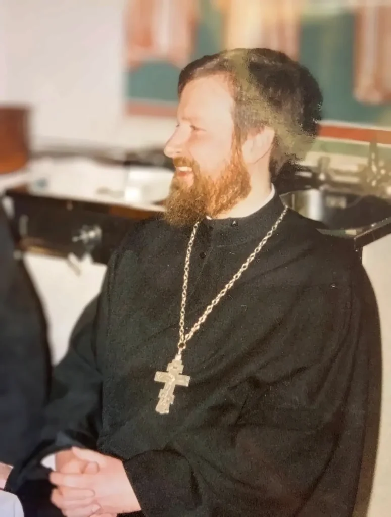 Der junge Vater Peter mit rotblondem Bart lacht im Profil, schwarzes Gewand, goldenes Brustkreuz an langer Kette, die Hände gefaltet