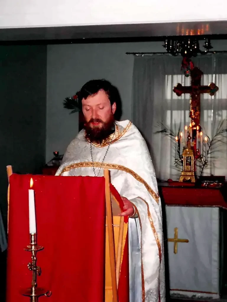 Vater Peter in weiß-goldenem Priestergewand am Lesepult mit rotem Tuch und brennender Kerze, im Hintergrund ein Kreuz und ein Leuchter mit Kerzen