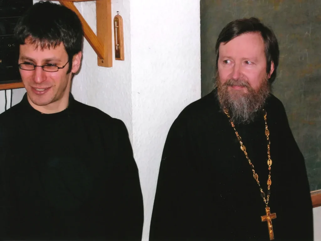Links Vater Vladimir, damals Diakon, mit Brille, rechts Vater Peter mit Bart und goldenem Brustkreuz, beide in schwarzem Gewand vor heller Wand und grüner Tafel
